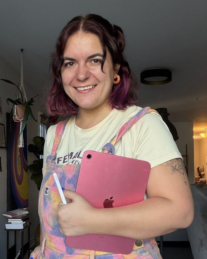 Anna smiling at home, holding a pink iPad and pencil, wearing pastel tie-dye overalls